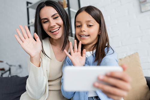 joyful babysitter and girl waving hands during video call on blurred smartphone