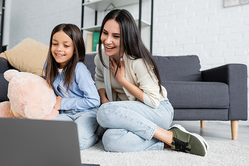 cheerful girl and nanny watching movie on laptop while sitting on floor