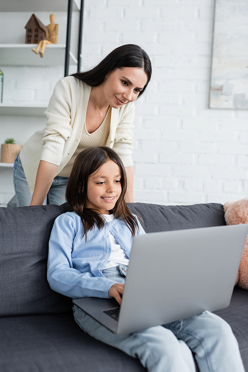 smiling babysitter looking at girl playing video game on laptop at home