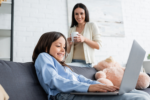excited girl gaming on laptop near nanny with tea cup on blurred background