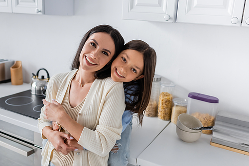happy babysitter with smiling child embracing and looking at camera in kitchen