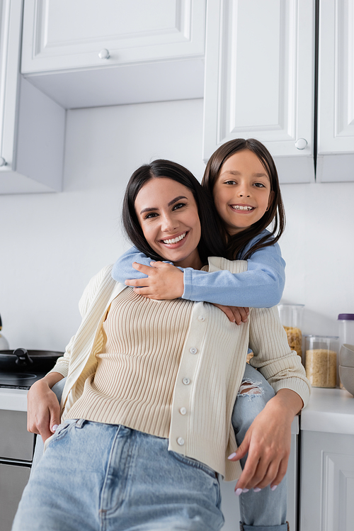 cheerful girl hugging happy nanny and smiling at camera in kitchen