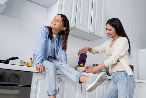 nanny pouring corn flakes near cheerful girl sitting on worktop in kitchen