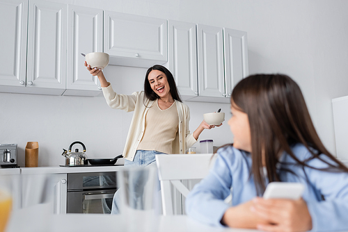 excited babysitter holding bowls with breakfast near blurred girl in kitchen