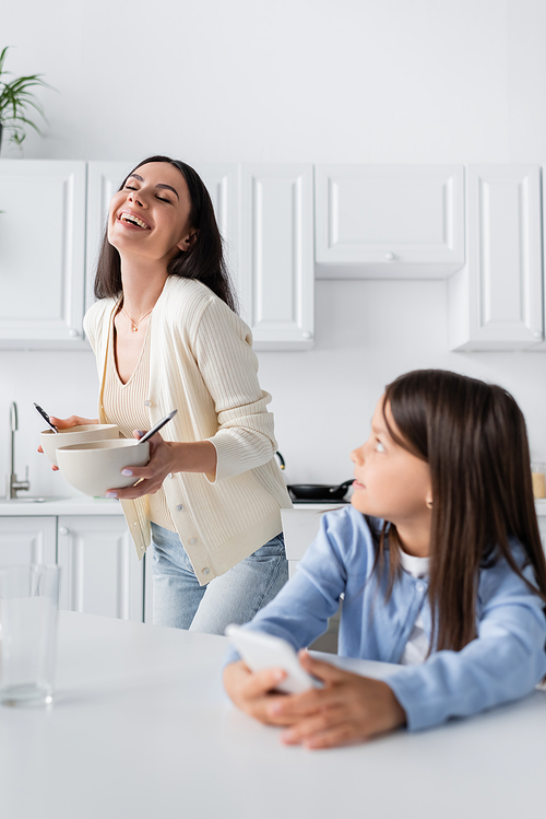 excited babysitter holding bowls with breakfast and laughing near girl in kitchen