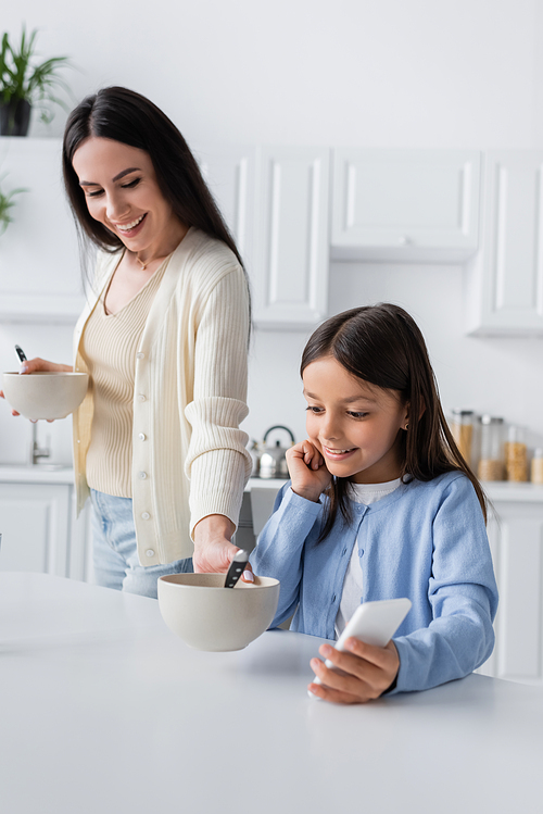 cheerful babysitter serving breakfast near girl with smartphone in kitchen