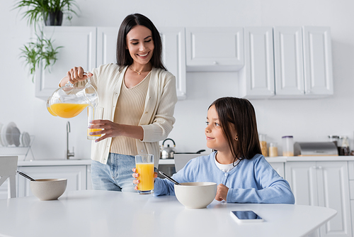 brunette nanny pouring orange juice near smiling girl having breakfast in kitchen