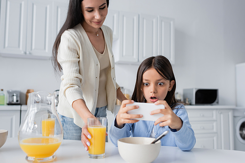 amazed girl looking at mobile phone near smiling nanny serving orange juice for breakfast