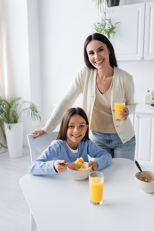 happy girl and nanny looking at camera near tasty corn flakes and fresh orange juice