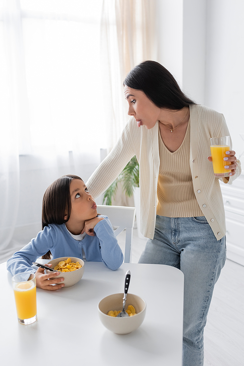 nanny and girl looking at each other and sticking out tongues during breakfast in kitchen