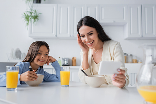 smiling nanny showing smartphone while having breakfast with girl in kitchen