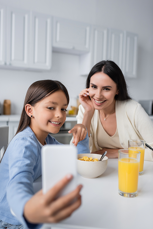 happy girl showing blurred mobile phone to babysitter during breakfast
