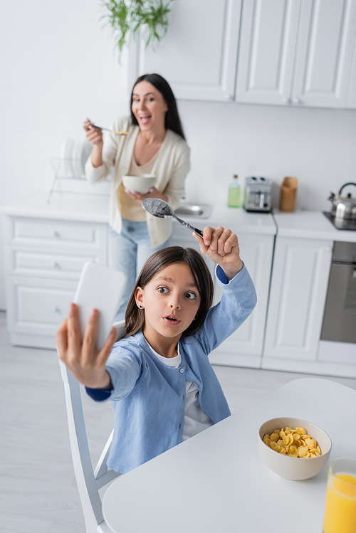 excited girl with spoon taking selfie near corn flakes and cheerful nanny on blurred background