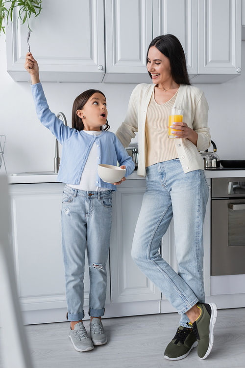 astonished girl holding spoon in raised hand near nanny with glass of orange juice