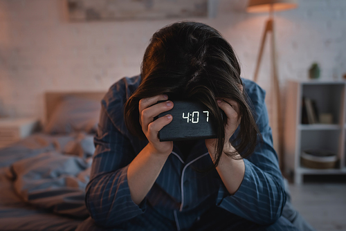 Young man with sleep disorder holding electronic clock on blurred bed at night