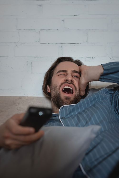 Tired man yawning and holding remote controller on bed at home