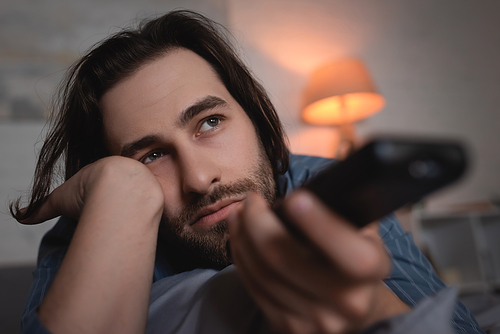 Brunette man with sleep disorder holding blurred remote controller in bedroom