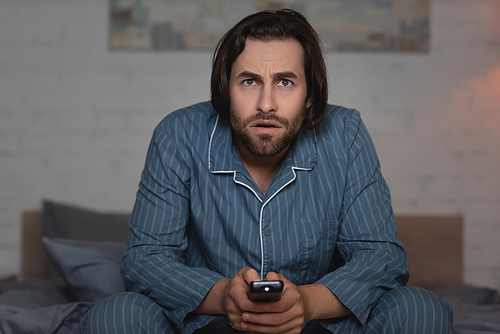 Man in pajama holding remote controller while watching tv on blurred bed