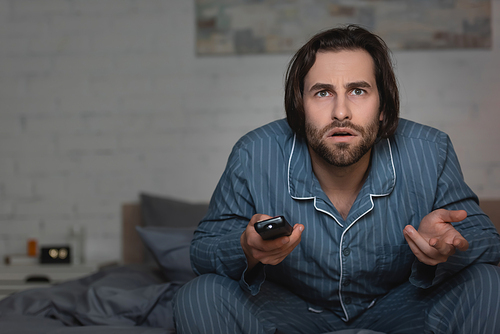 Confused man with insomnia holding remote controller on bed at night