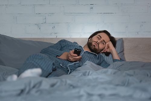 Tired man in pajama holding remote controller while lying on bed