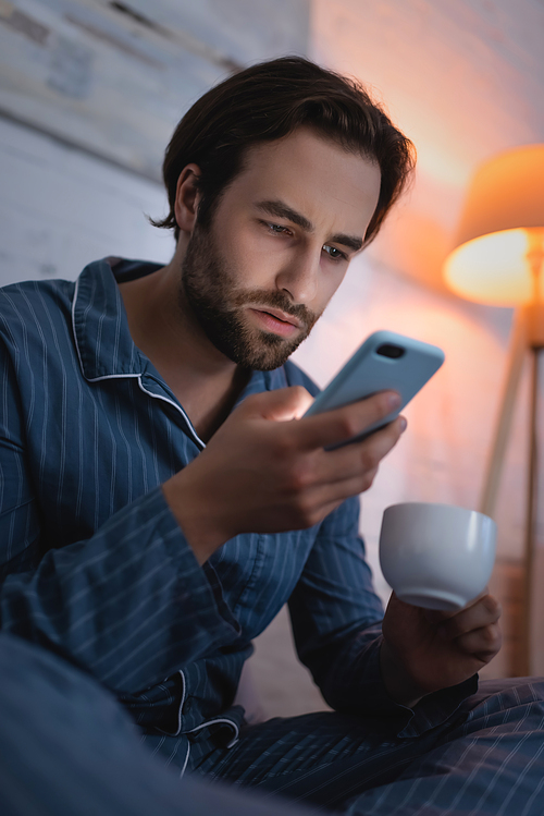 Young man holding cup of coffee and using smartphone in bedroom at night