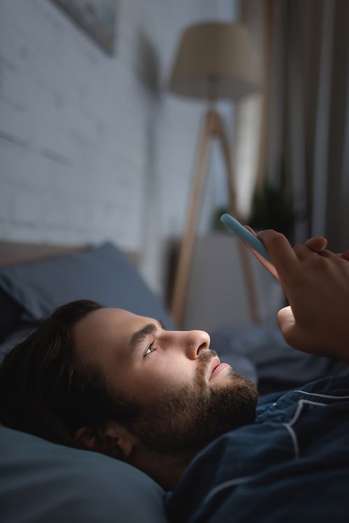 Side view of young man with sleep disorder using mobile phone on bed at night