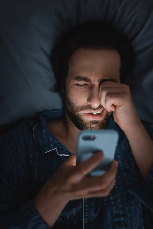 Top view of exhausted man in pajama using smartphone on bed at night