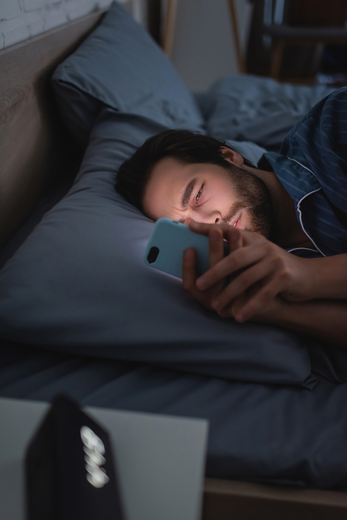 Sleepless man using mobile phone near blurred alarm clock on bed at night