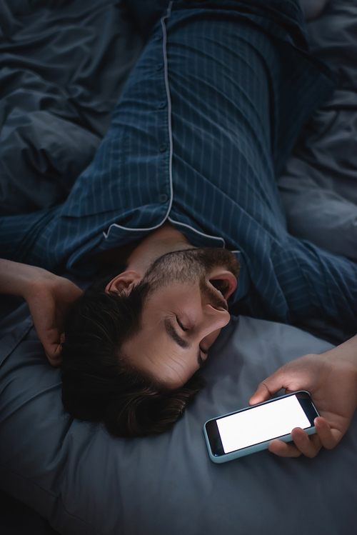 Top view of exhausted man yawning and holding smartphone on bed at home