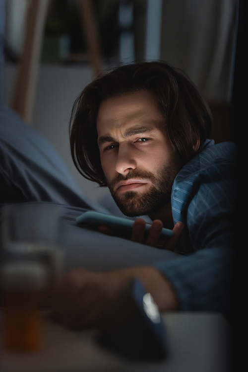 Dissatisfied man with insomnia holding smartphone on bed at home