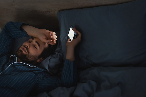 Young man with sleep disorder holding cellphone on bed in bedroom