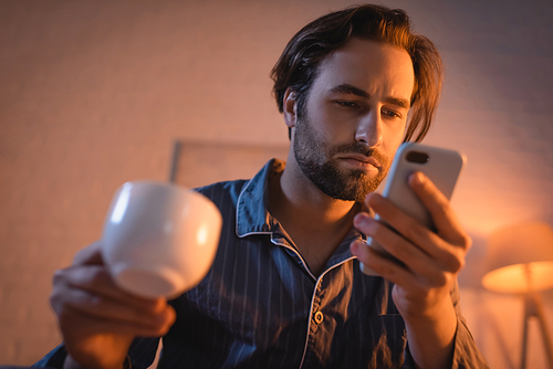 Young man in pajamas holding blurred coffee and smartphone at night