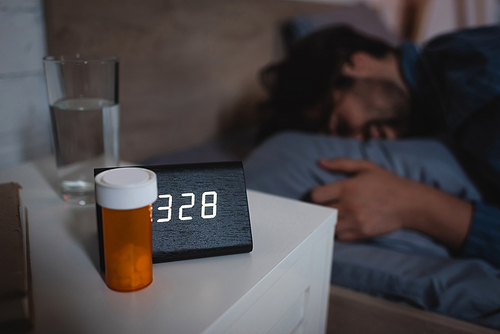 Pills and alarm clock on bedside table near blurred man on bed at night