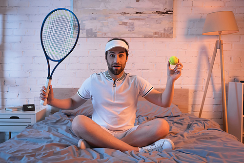 Amazed somnambulist in sportswear holding tennis ball and rocket on bed at night