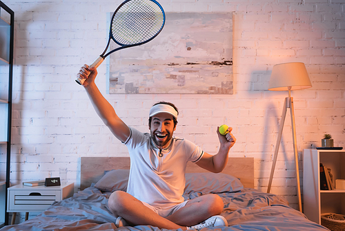 Cheerful sleepwalker in sportswear holding tennis rocket and ball on bed