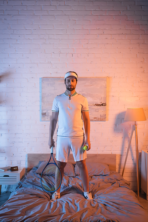 Sleepwalker in sportswear holding tennis rocket and ball on bed at home