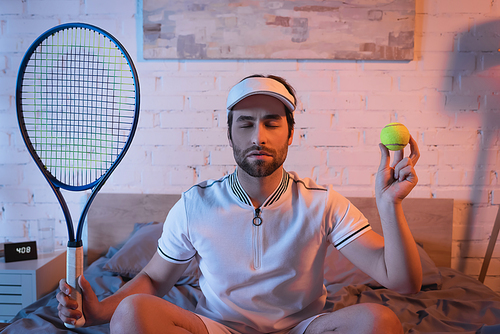 Sleepwalker with closed eyes holding tennis rocket and ball on bed