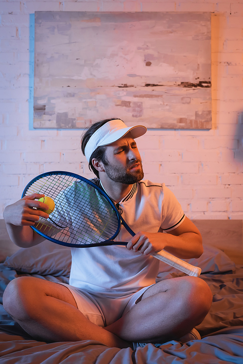 Confused sleepwalker holding tennis racket and ball while sitting on bed at night