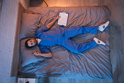 Top view of sleepwalker in doctor uniform holding clipboard and pen while sleeping on bed