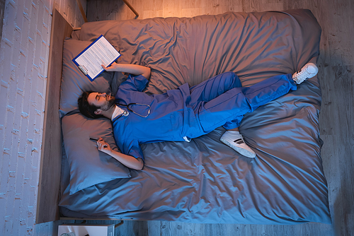 Young sleepwalker in doctor uniform holding pen and clipboard while sleeping on bed