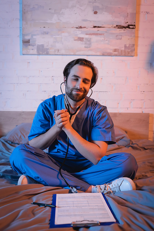 Smiling somnambulist in doctor uniform and stethoscope sitting near clipboard on bed