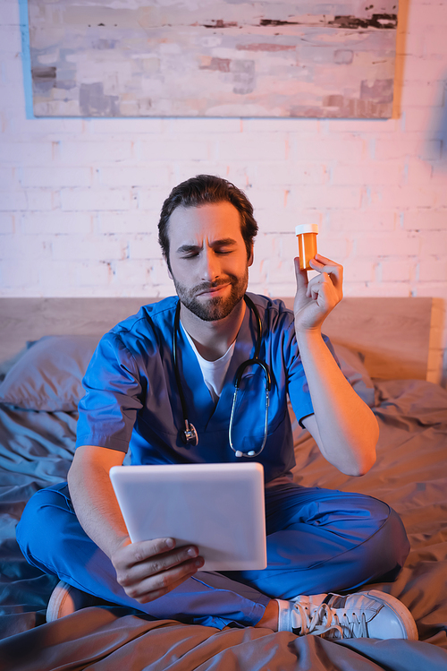 Confused doctor holding pills and digital tablet while suffering from sleepwalking on bed