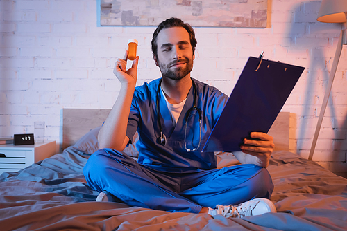 Smiling sleepwalker in doctor uniform holding clipboard and pills on bed at night