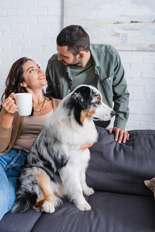smiling young woman holding cup and looking at bearded boyfriend near australian shepherd dog