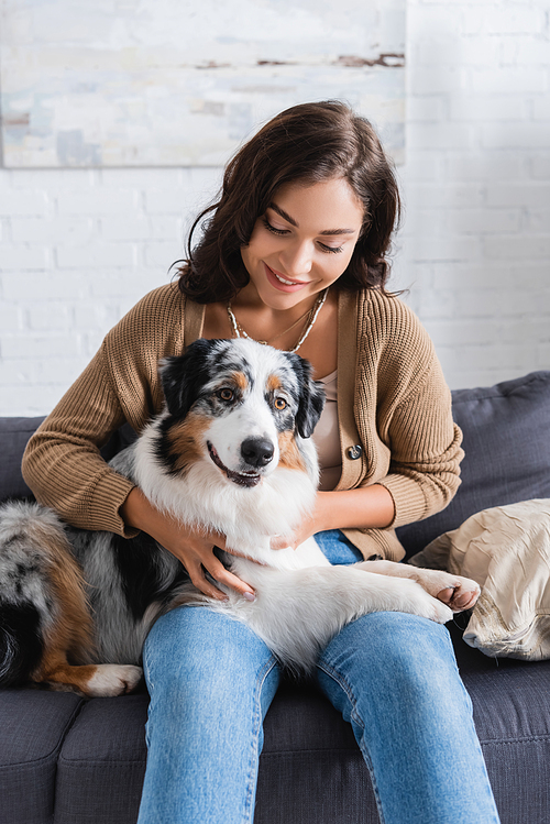 smiling young woman cuddling australian shepherd dog while sitting on couch