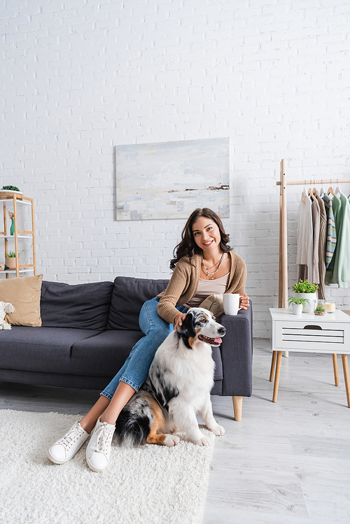 happy young woman sitting on sofa and cuddling australian shepherd dog while holding cup of coffee
