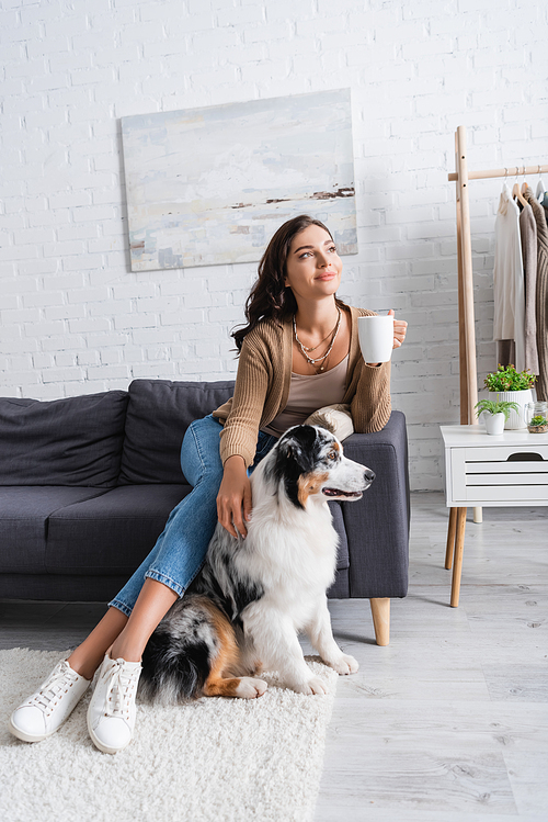 smiling young woman sitting on sofa and cuddling australian shepherd dog while holding cup of coffee
