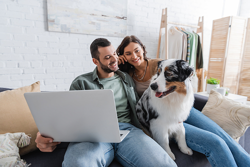 happy young couple with laptop looking at australian shepherd dog