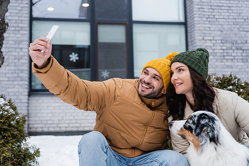 happy man in knitted hat taking selfie with girlfriend and australian shepherd dog in winter
