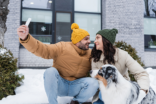 happy man in knitted hat taking selfie with cheerful girlfriend and australian shepherd dog in winter